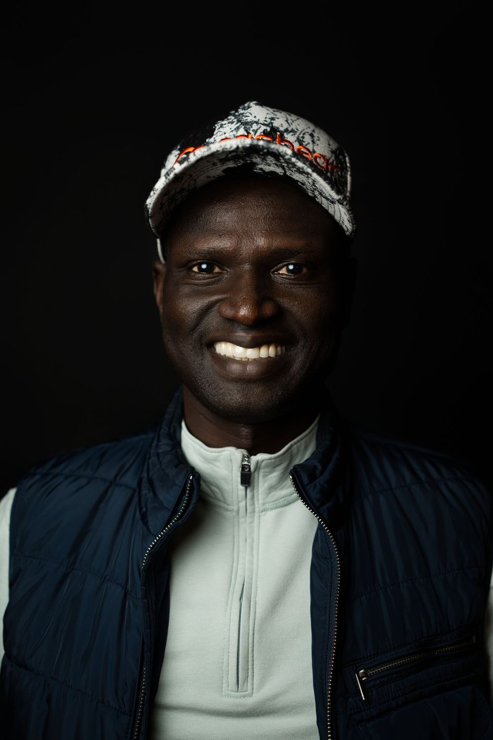 Portrait of someone wearing a blue jacket and camouflage cap against a dark background with a bright smile.