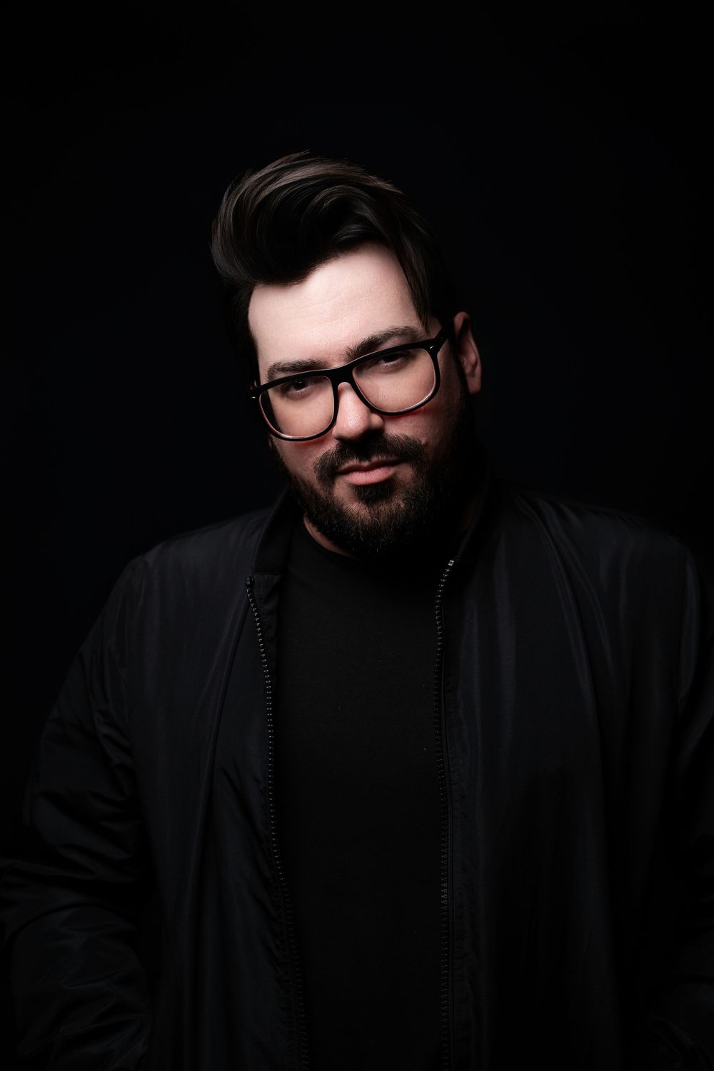 A dark studio portrait shows a person with a beard and glasses wearing black clothing against a black background.