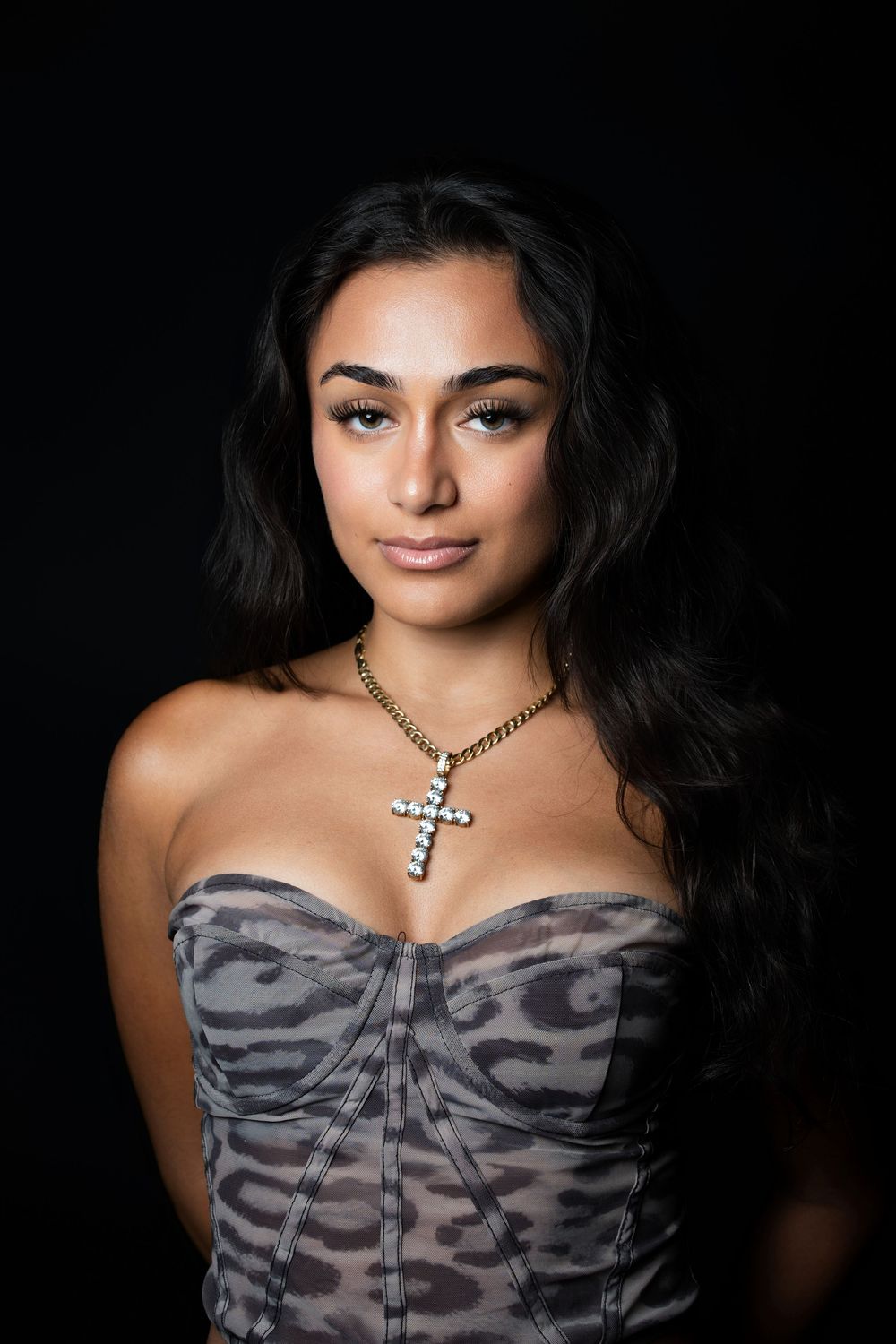 A figure in a gray strapless top with dark wavy hair and a cross necklace poses against a black background.