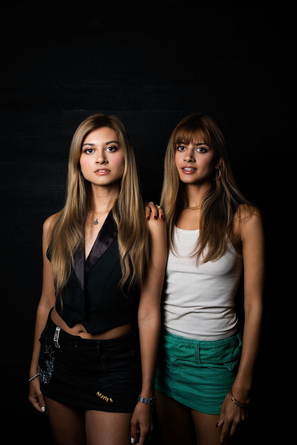 Two models pose together in contrasting black and white outfits with emerald green shorts against dark backdrop.