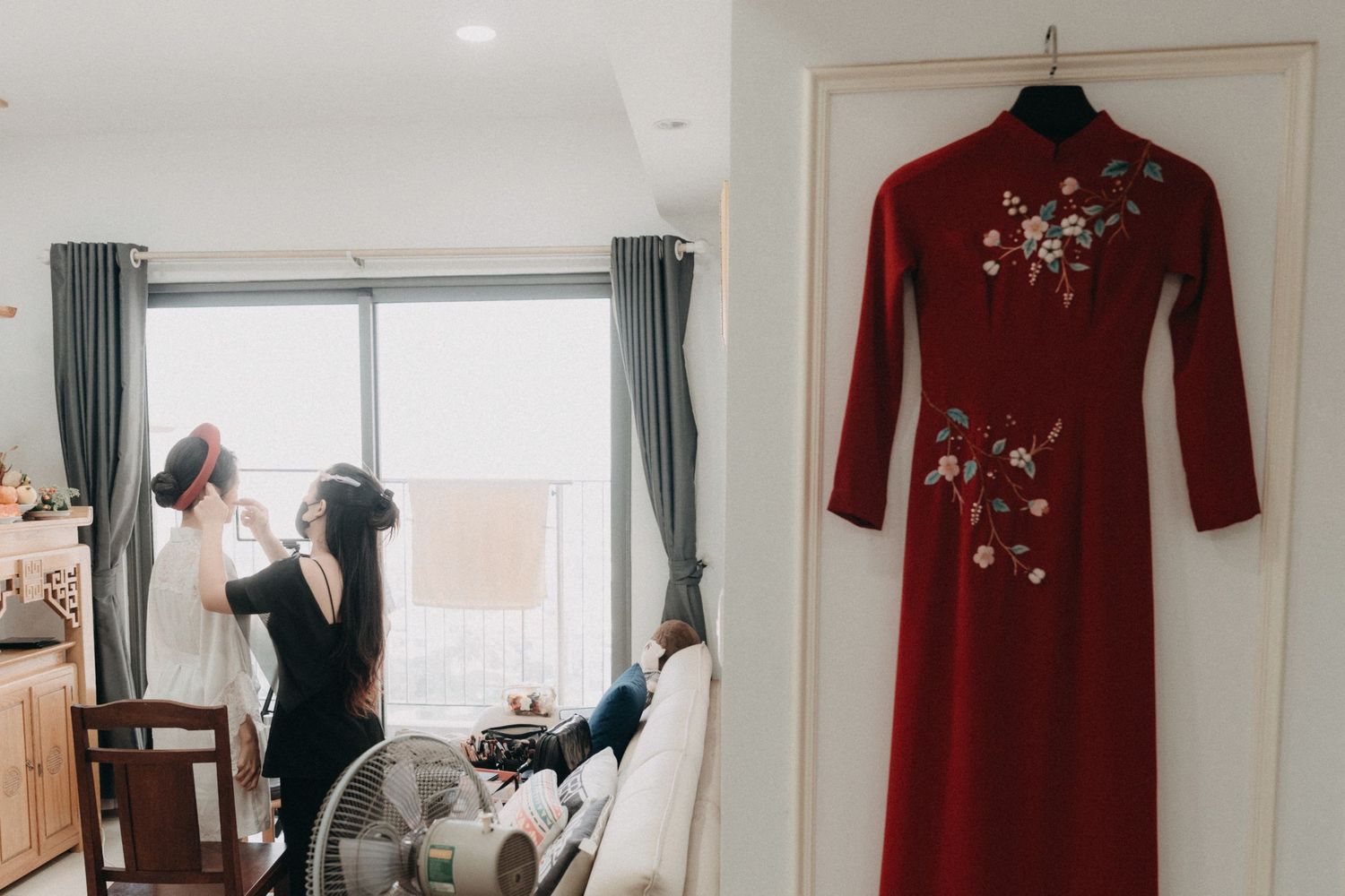 Red embroidered Vietnamese ao dai hanging beside bride getting ready in bright room.