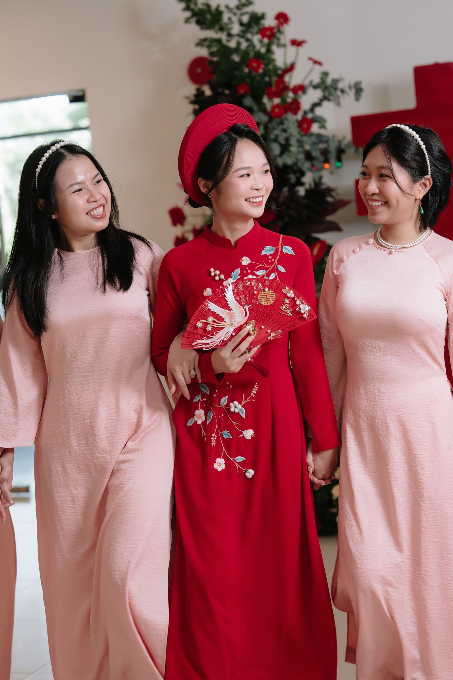 Vietnamese bride in red ao dai and hat walks with two smiling bridesmaids in pink ao dai.