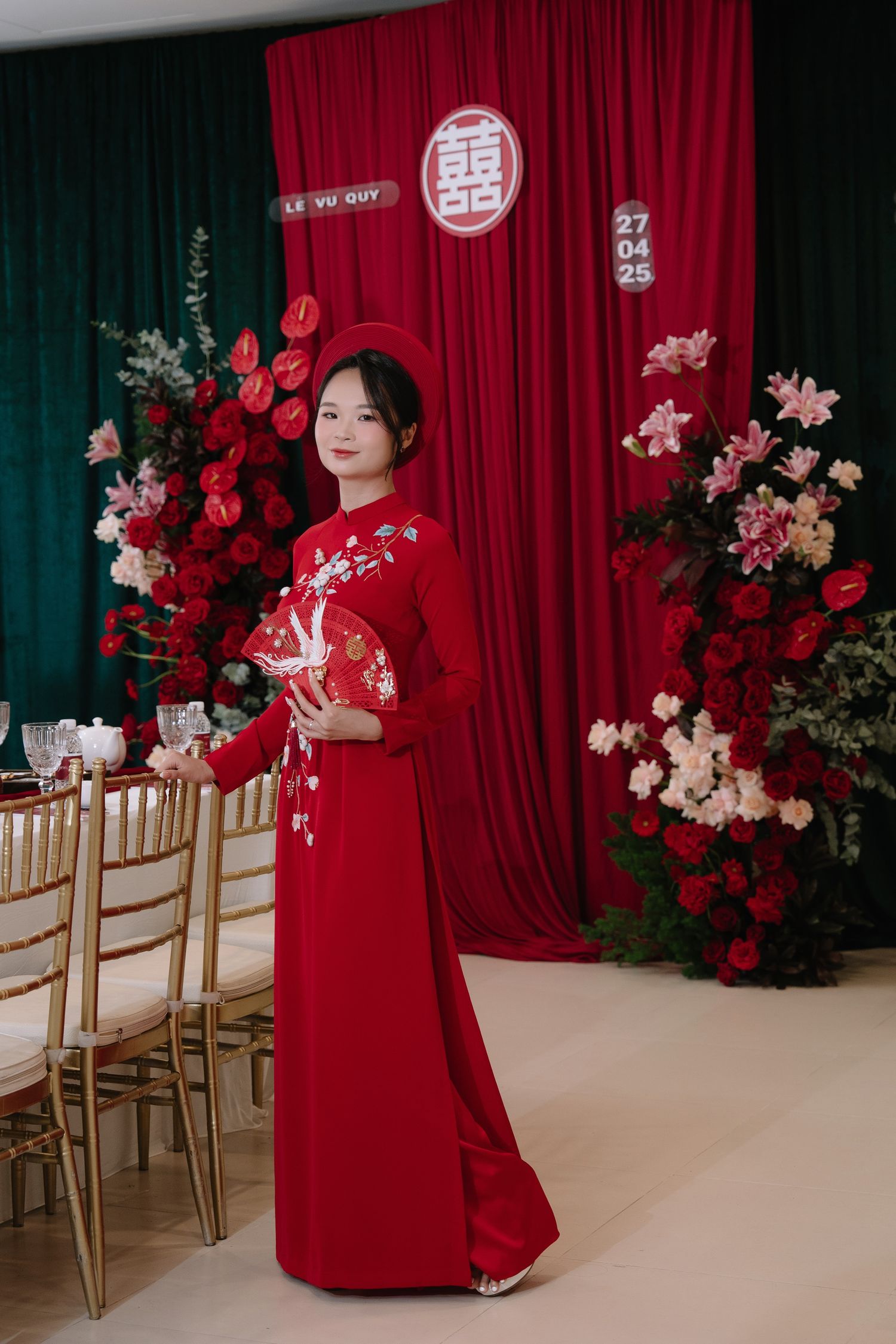 Vietnamese bride in embroidered red ao dai holds decorative fan at traditional Le Vu Quy wedding ceremony.