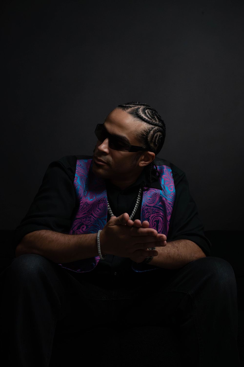 Man with cornrow braids sits in dark studio, hands clasped, wearing black outfit with colorful paisley vest and chain.