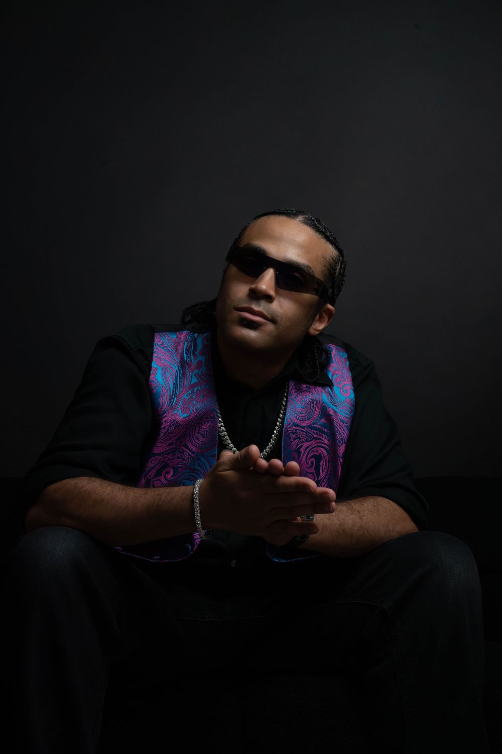 Man in sunglasses sits in dark studio with hands clasped, wearing black shirt and vibrant blue-pink paisley vest.