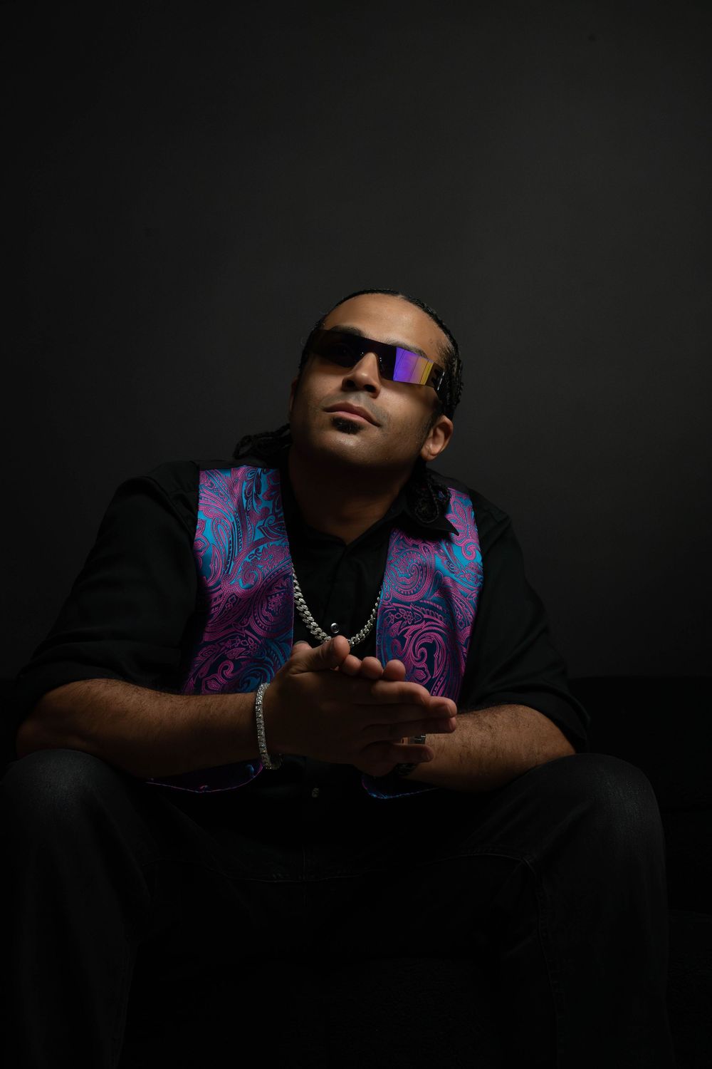 Man in purple-tinted sunglasses poses in dark studio wearing black outfit with colorful paisley vest and silver chain.