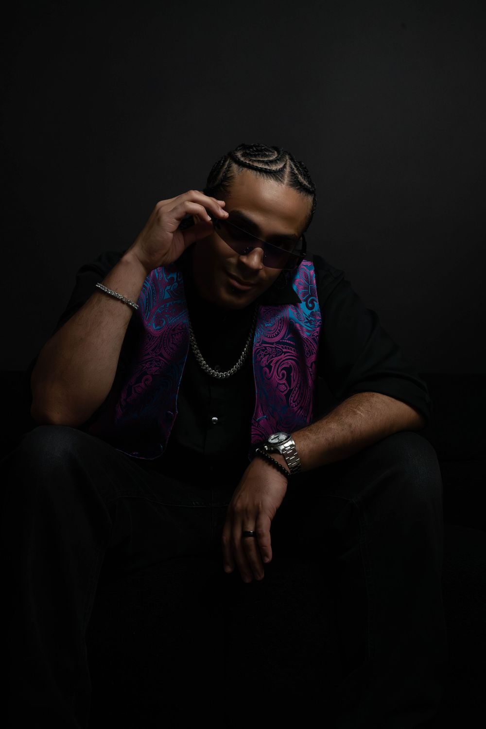 Man in sunglasses adjusts frames while seated in dark studio, wearing black outfit with bold paisley vest and watch.