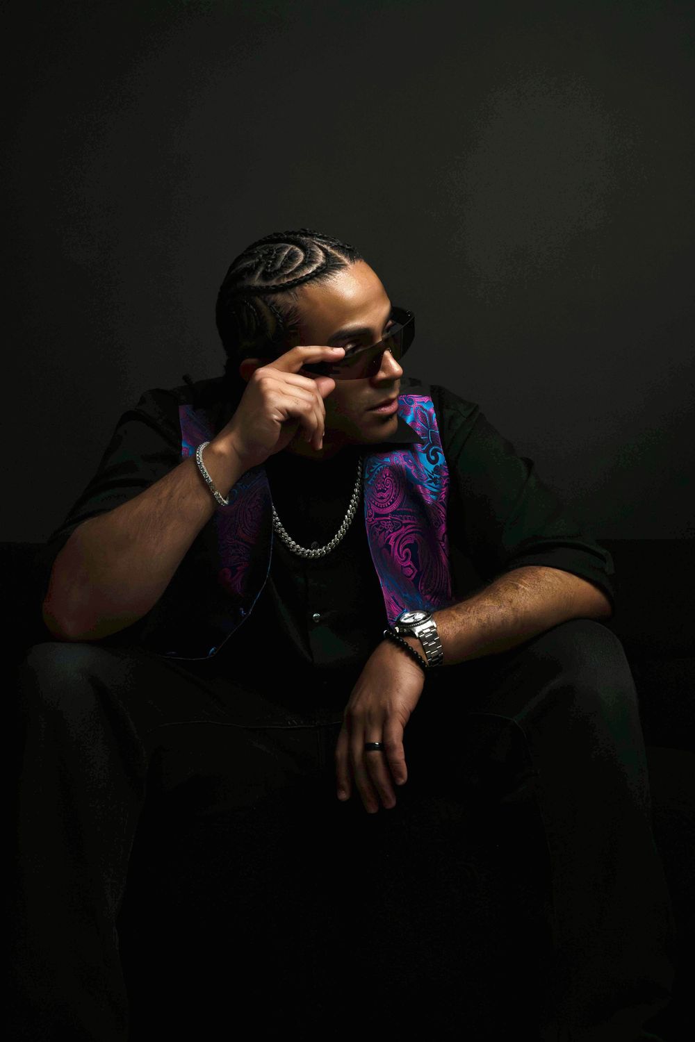 Man with cornrow braids points to temple in dark studio, wearing sunglasses, paisley vest, and silver chain necklace.