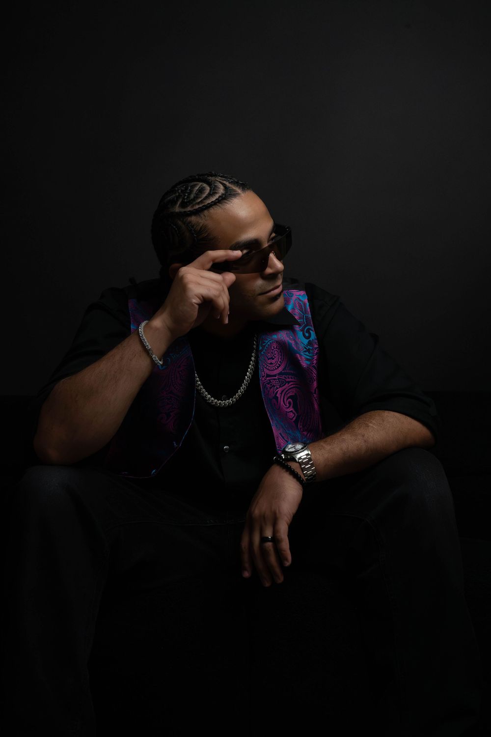 Man in sunglasses gestures to temple while seated in dark studio, wearing black outfit with colorful paisley vest.