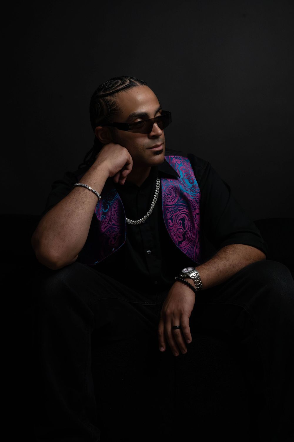 Man in sunglasses rests chin on hand in dark studio, wearing black shirt with vibrant paisley vest and silver chain.