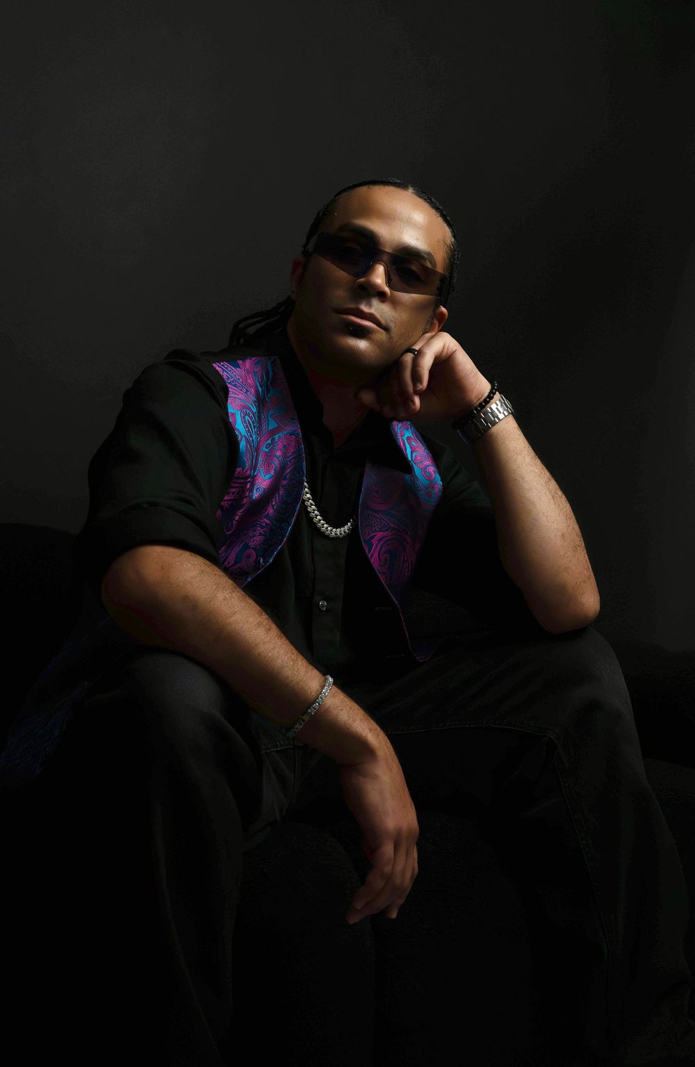 Man in sunglasses rests chin on hand in dark studio, wearing black outfit with blue-pink paisley vest and bracelet.
