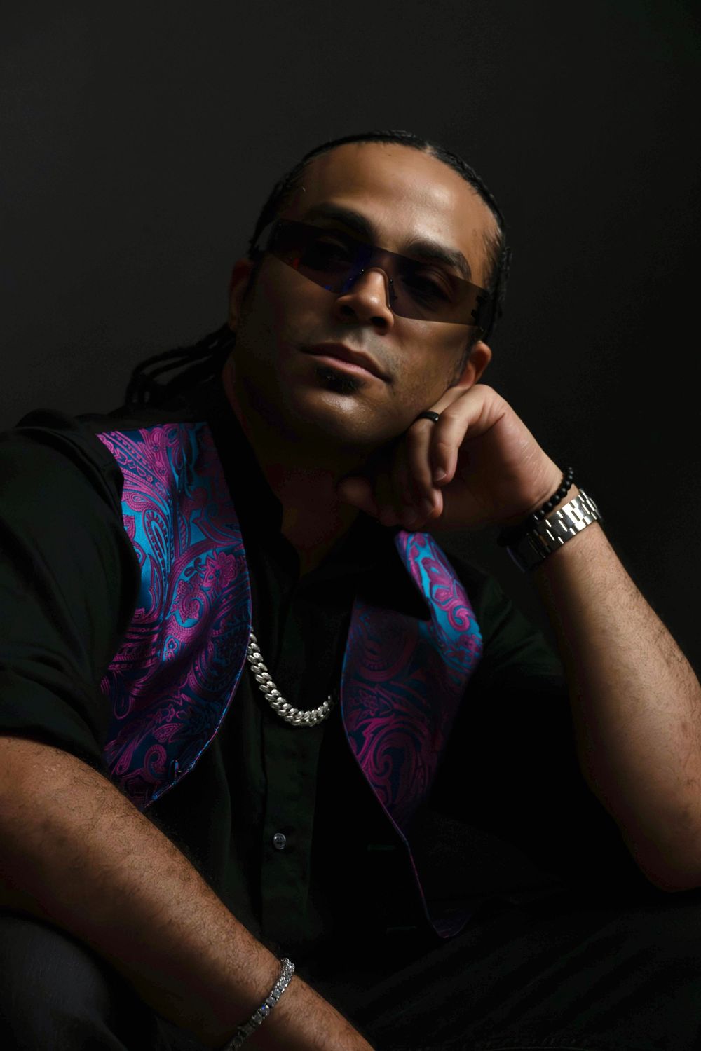 Man in sunglasses rests chin on hand in dark studio, wearing black shirt with colorful paisley vest and silver chain.