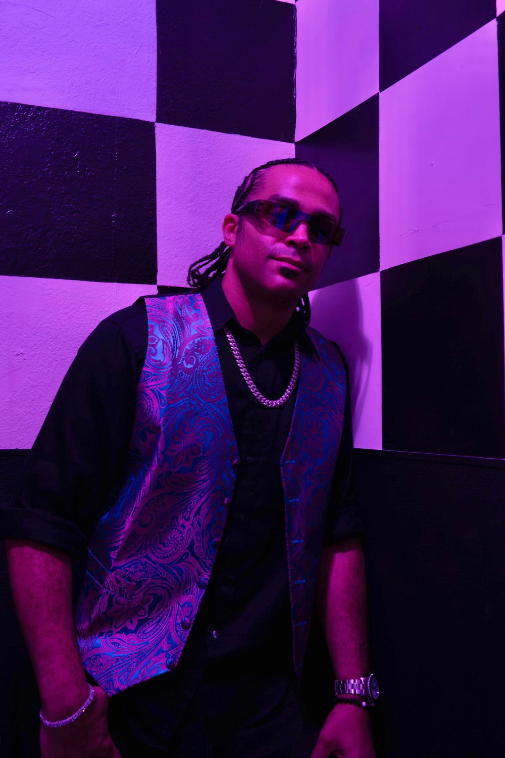 Man in sunglasses leans against checkered wall under purple neon light, wearing black outfit with paisley vest.