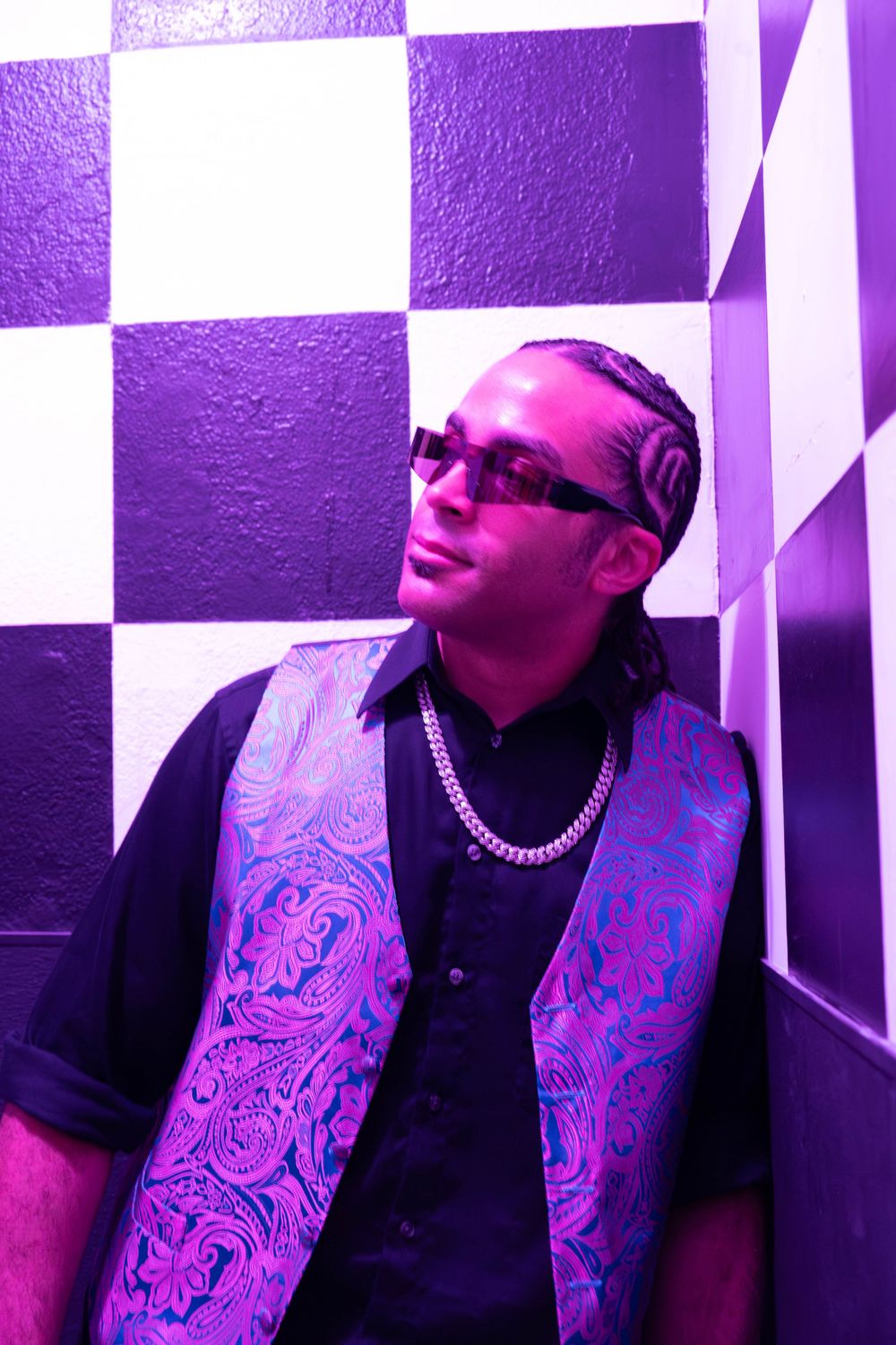 Man in purple paisley vest and sunglasses poses against checkered wall in purple neon light.