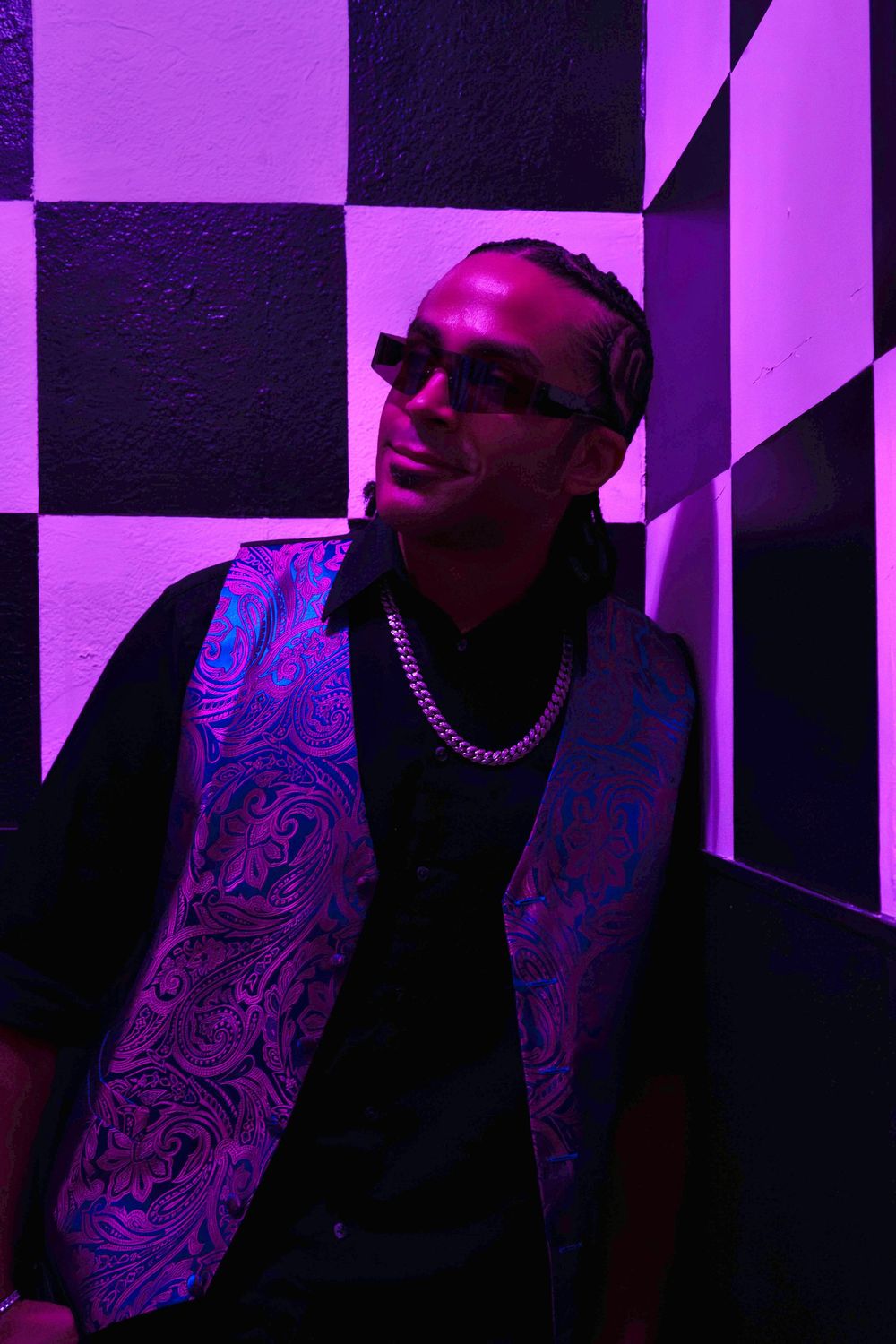 Man in decorative purple vest and sunglasses smiles under purple neon lighting against checkered wall.