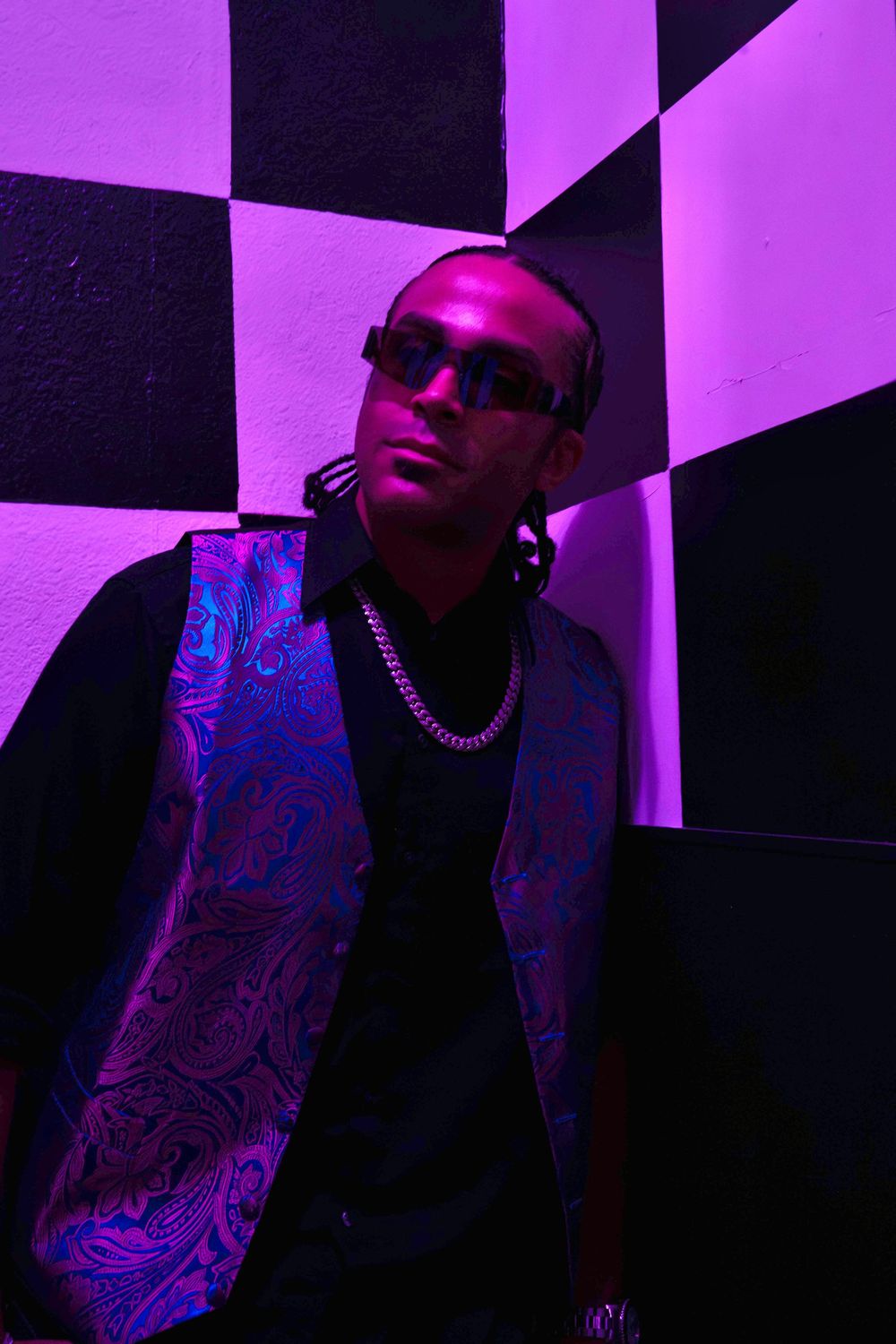 Stylish man in paisley vest and shades leans against black and white checkered wall in purple light.