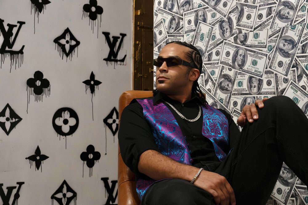 Man in colorful paisley vest sits relaxed in chair between Louis Vuitton and money print backdrops.