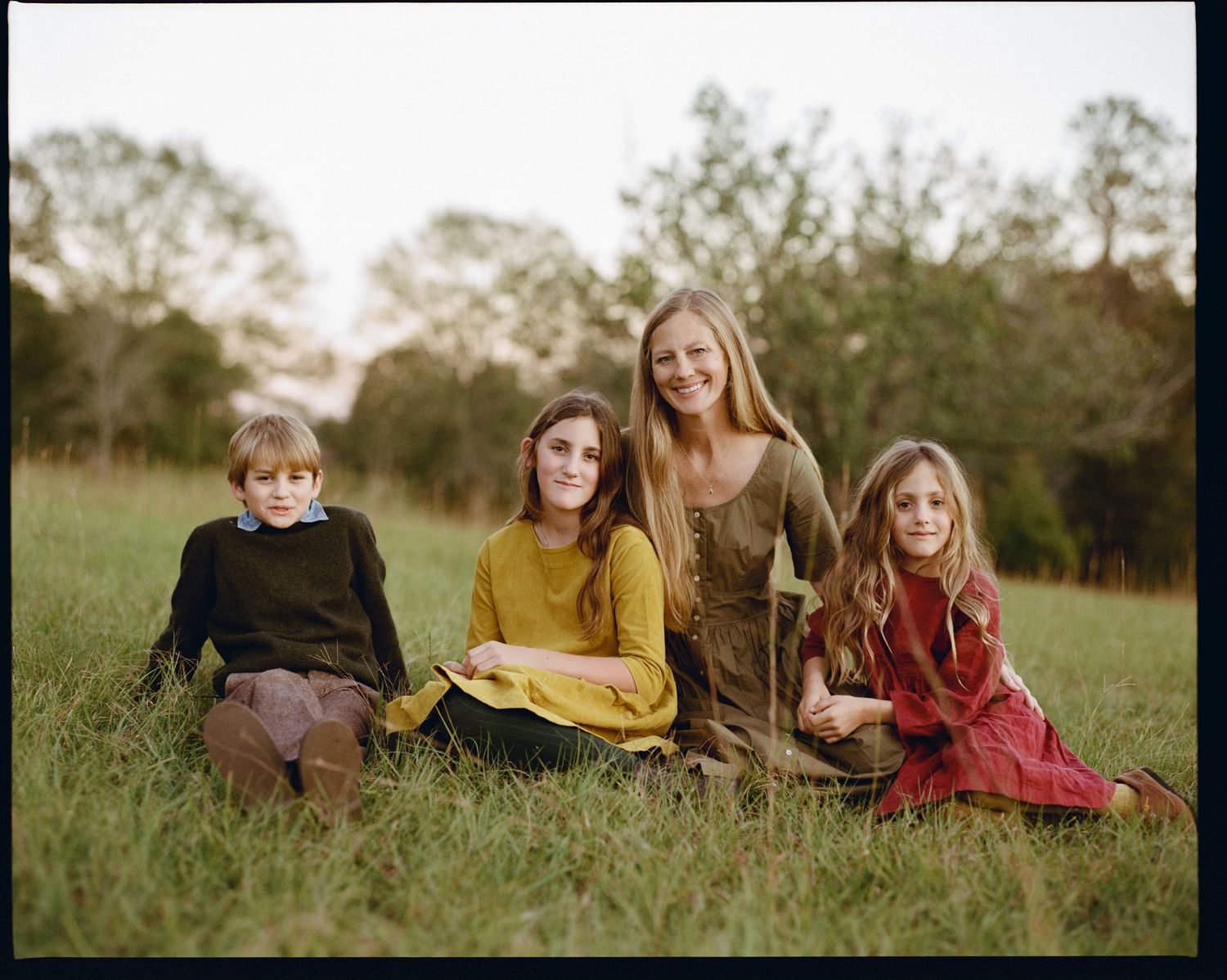 Family wearing coordinating fall clothing poses casually on grassy field at sunset.