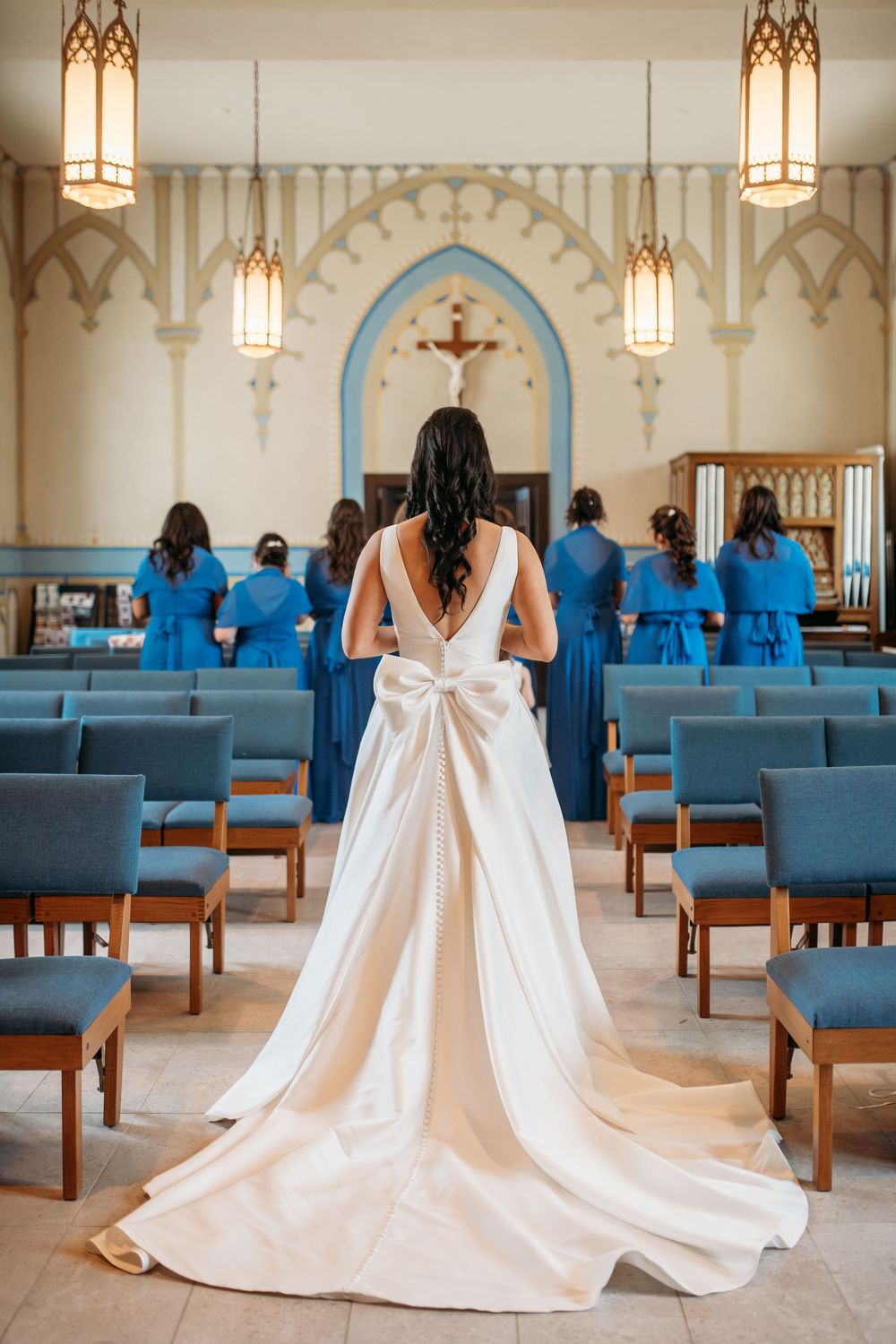 A bride in a flowing white gown with a dramatic bow stands in a chapel before bridesmaids wearing bright blue dresses.