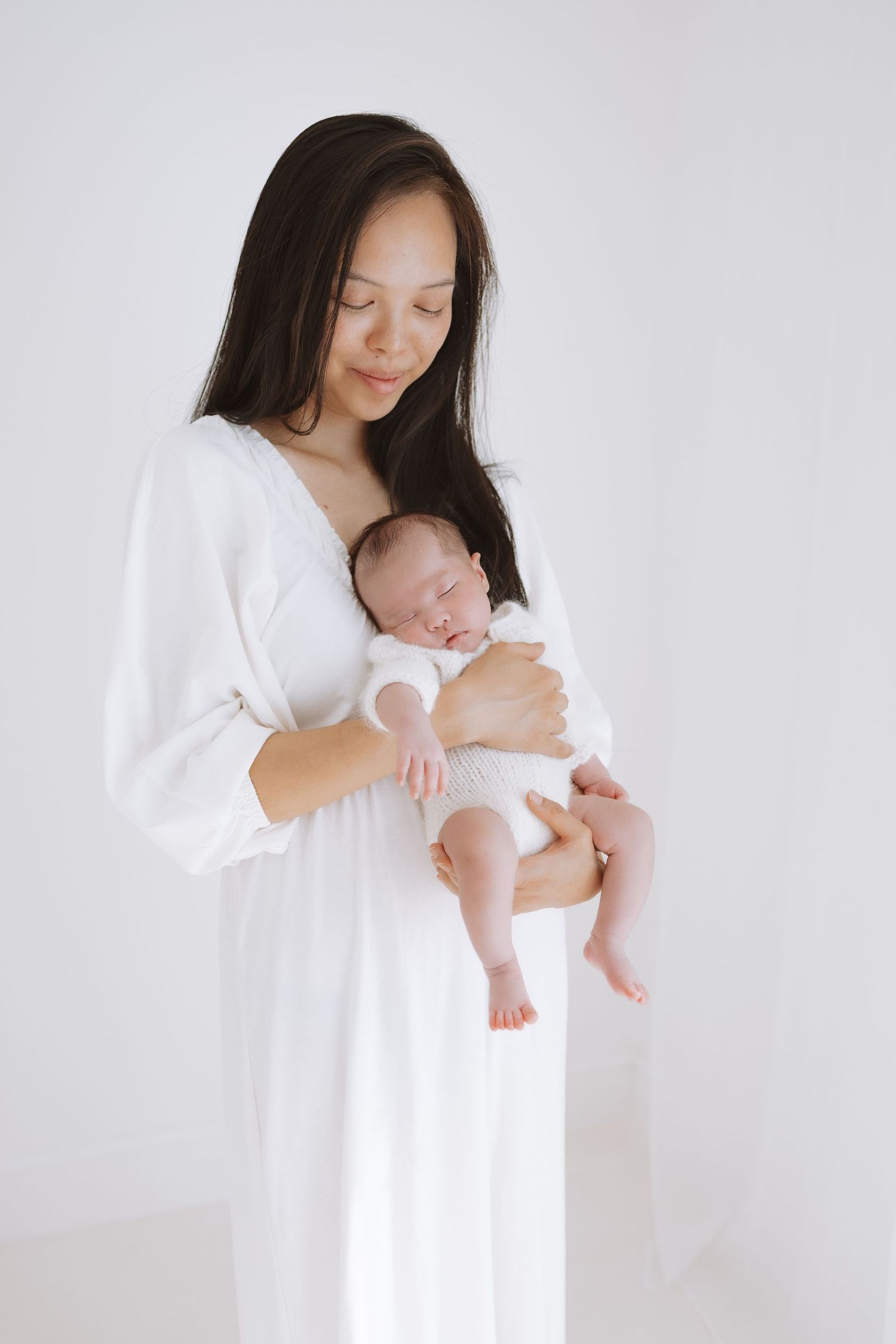 A mother in a white dress holds her sleeping newborn baby against her chest in a minimalist studio setting.