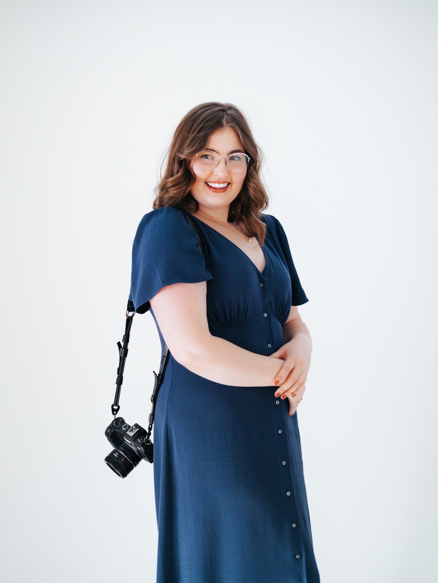 Female photographer in navy dress poses with camera on white background.