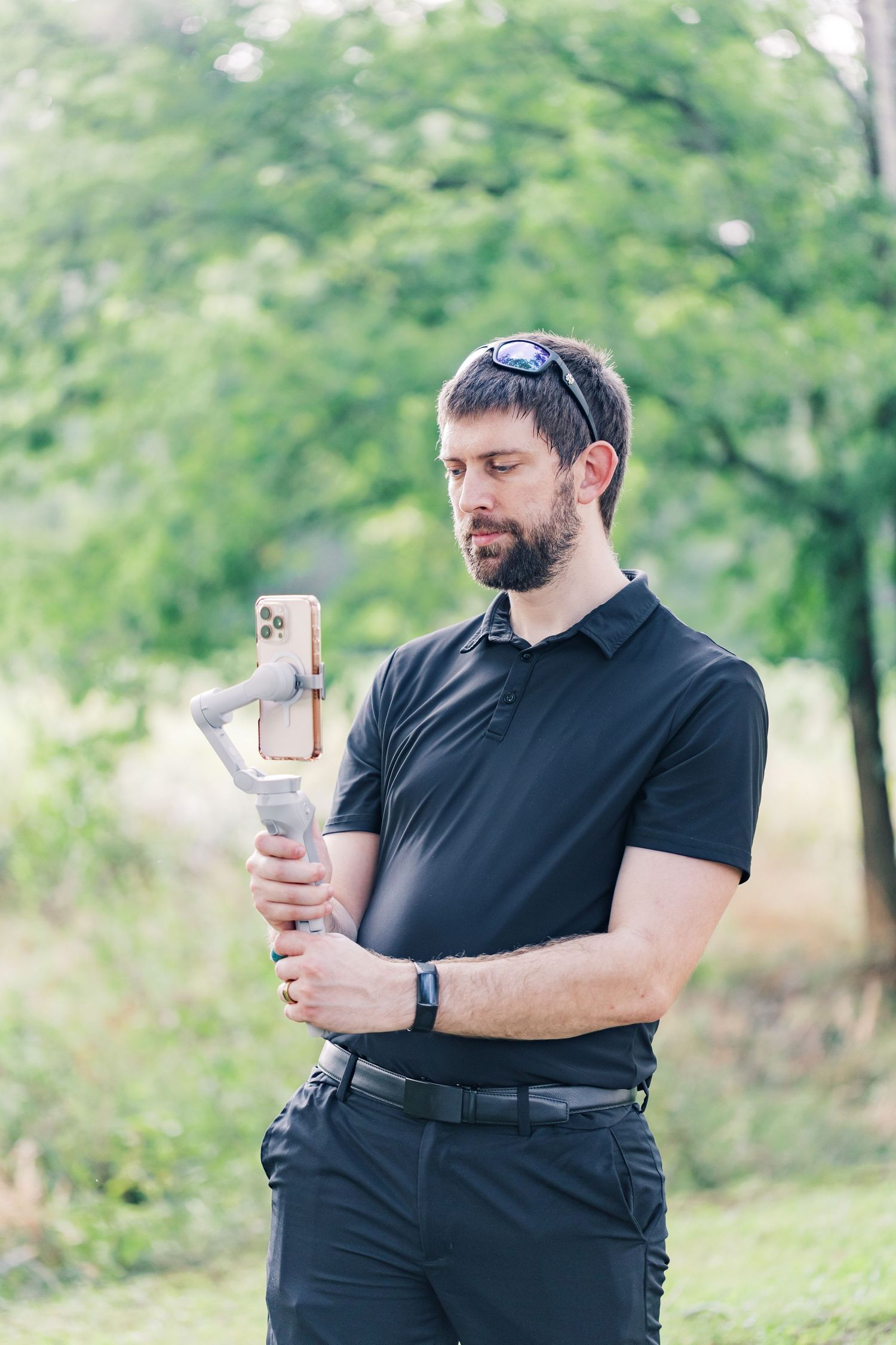 Bearded male videographer holds a smartphone gimbal stabilizer outdoors in a green park.