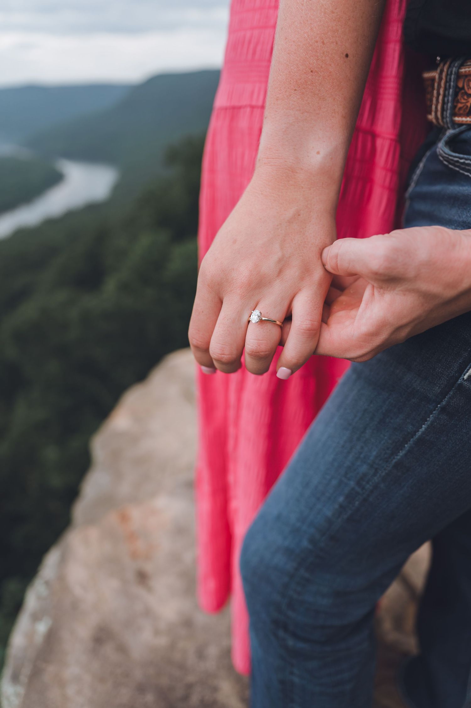 A couple holds hands showing an engagement ring with a scenic mountain view and winding river in the background.