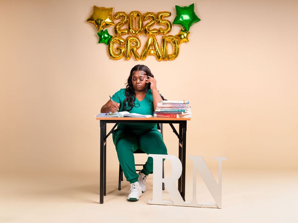 Graduate in green dress sits at desk with scattered papers and gold celebration balloons in studio setting.