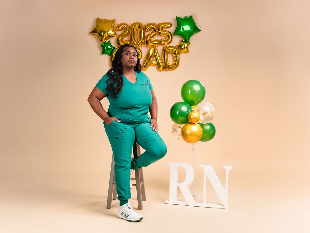 Nursing graduate in teal scrubs posing with celebratory balloons and decorations.