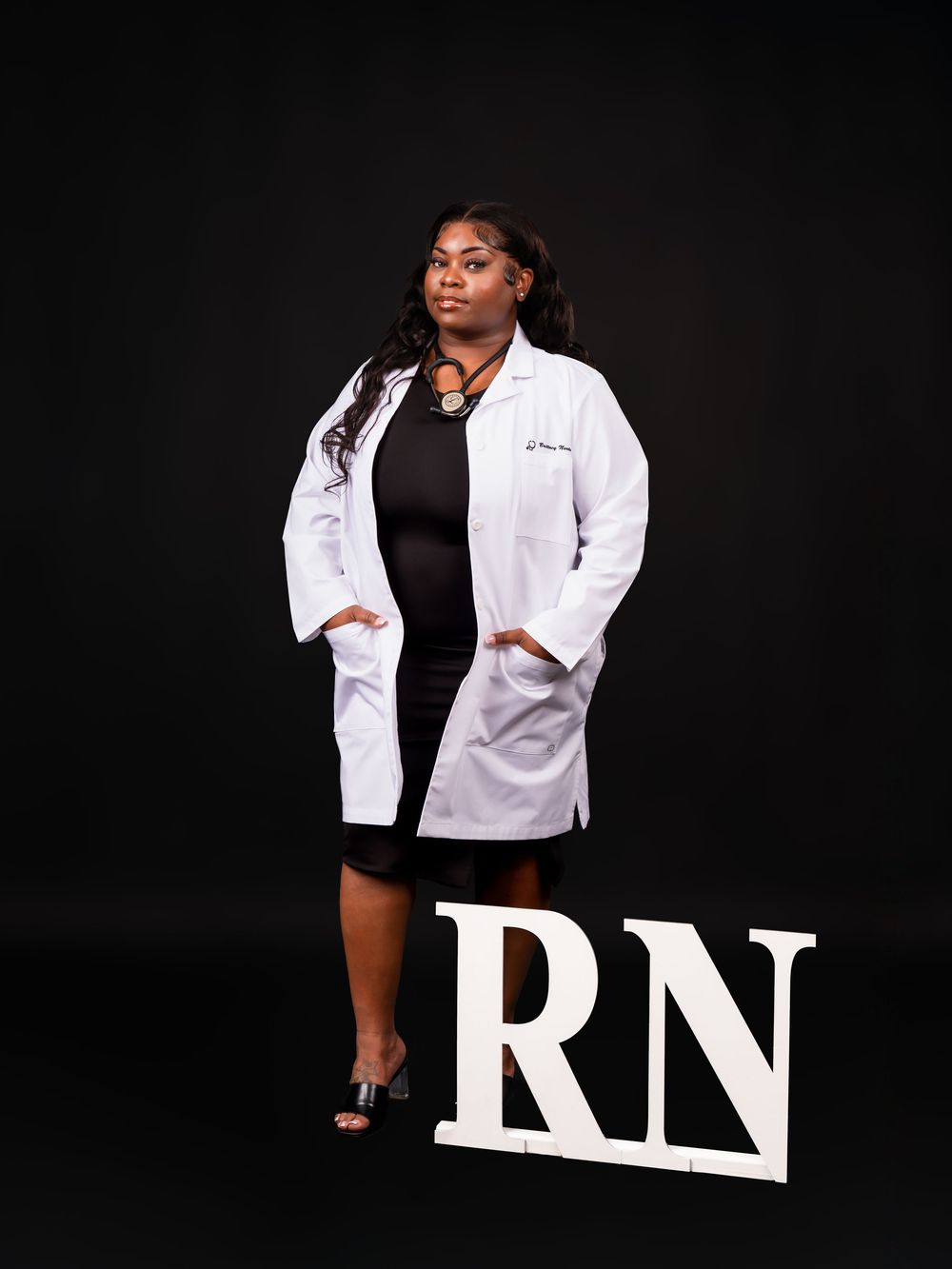 Professional wearing white lab coat poses with RN letters in dark studio setting.