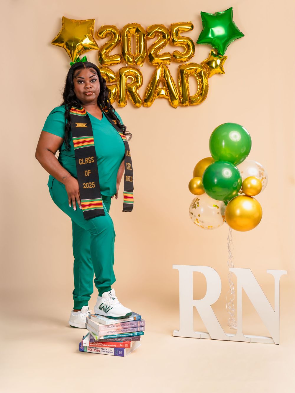 Graduate in teal scrubs with kente stole posing with books and balloon decorations.