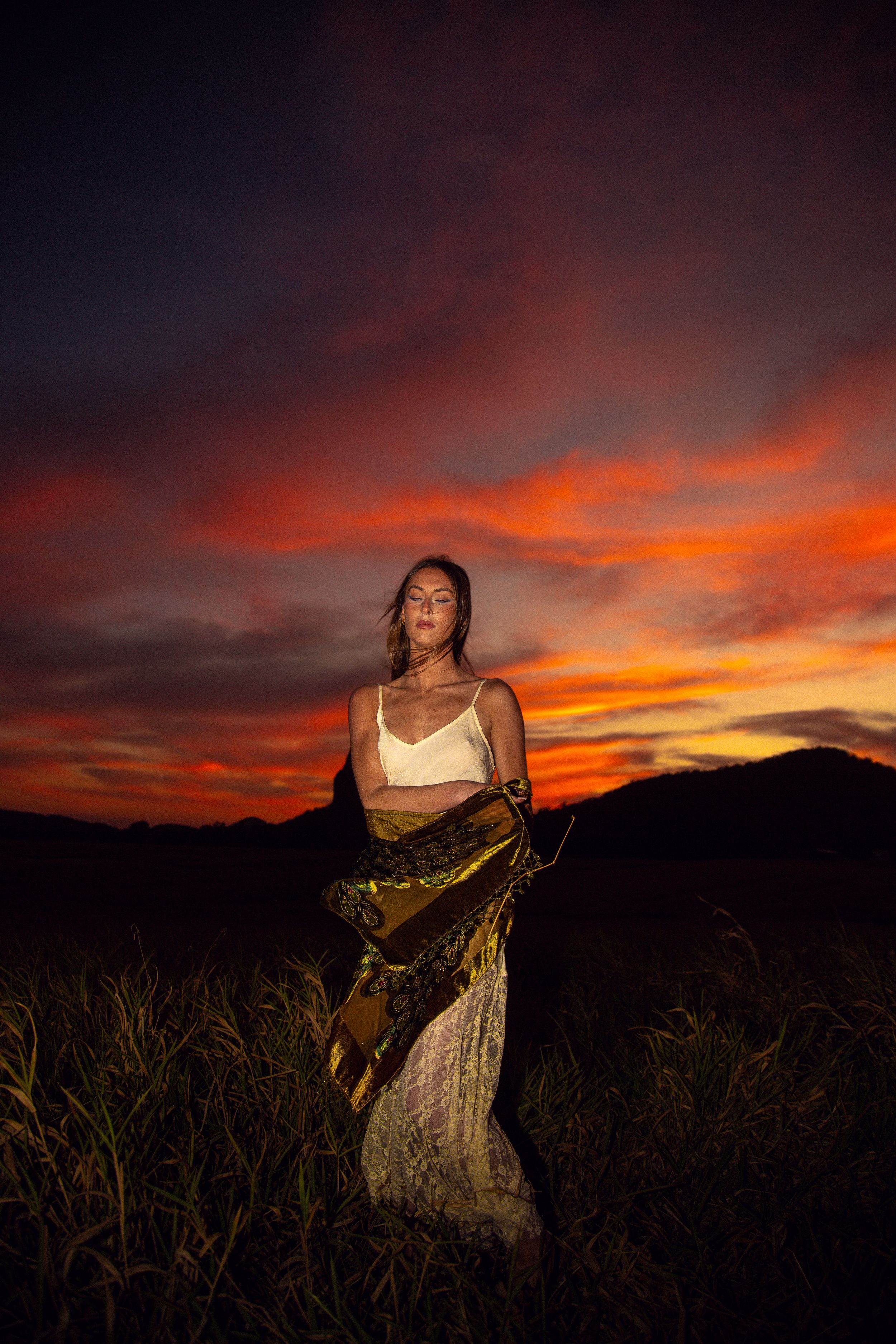 A figure in a flowing dress poses dramatically against a vibrant orange and purple sunset sky over mountain silhouettes.