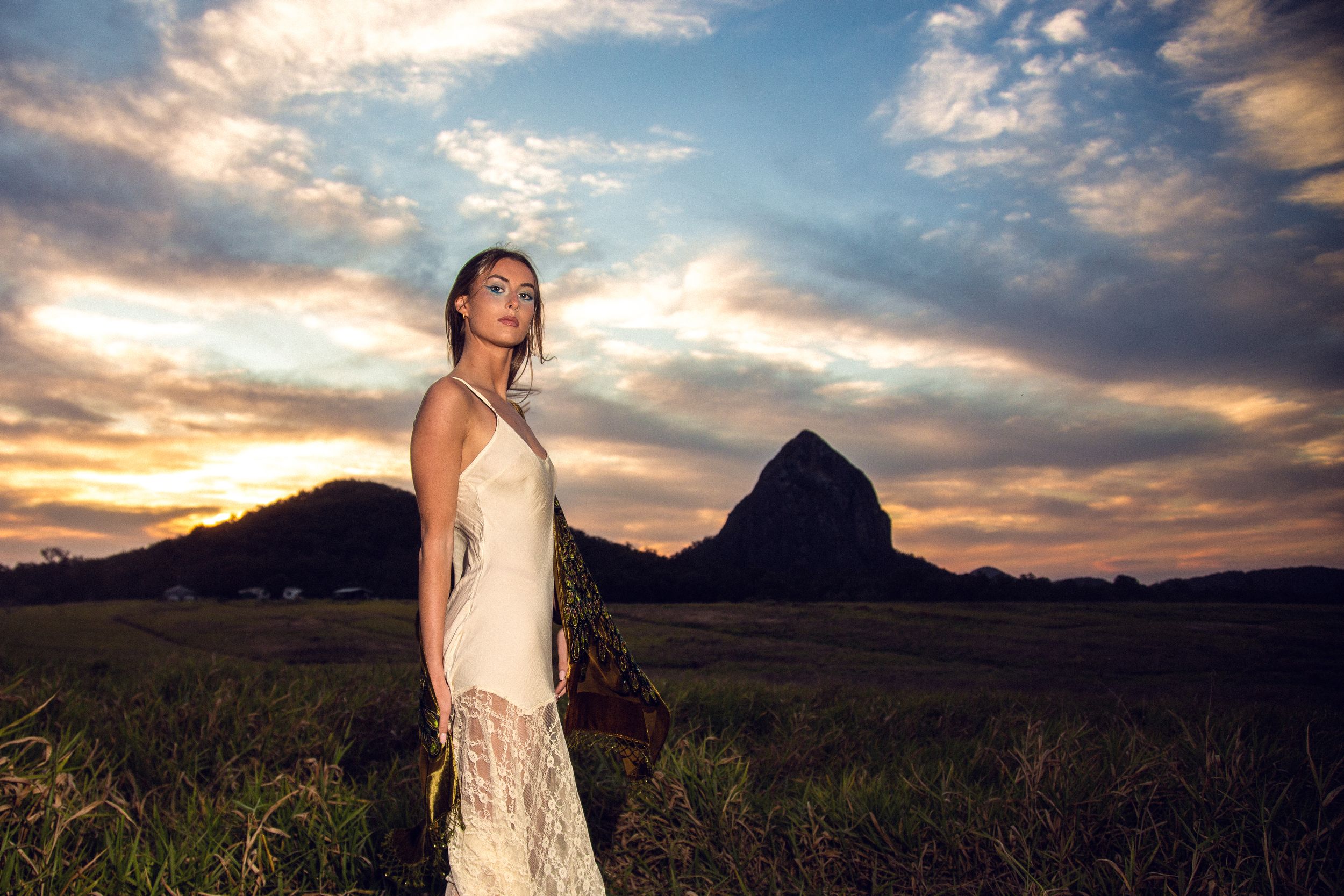 A figure stands in a field against a dramatic sunset sky with clouds and mountains in the background.