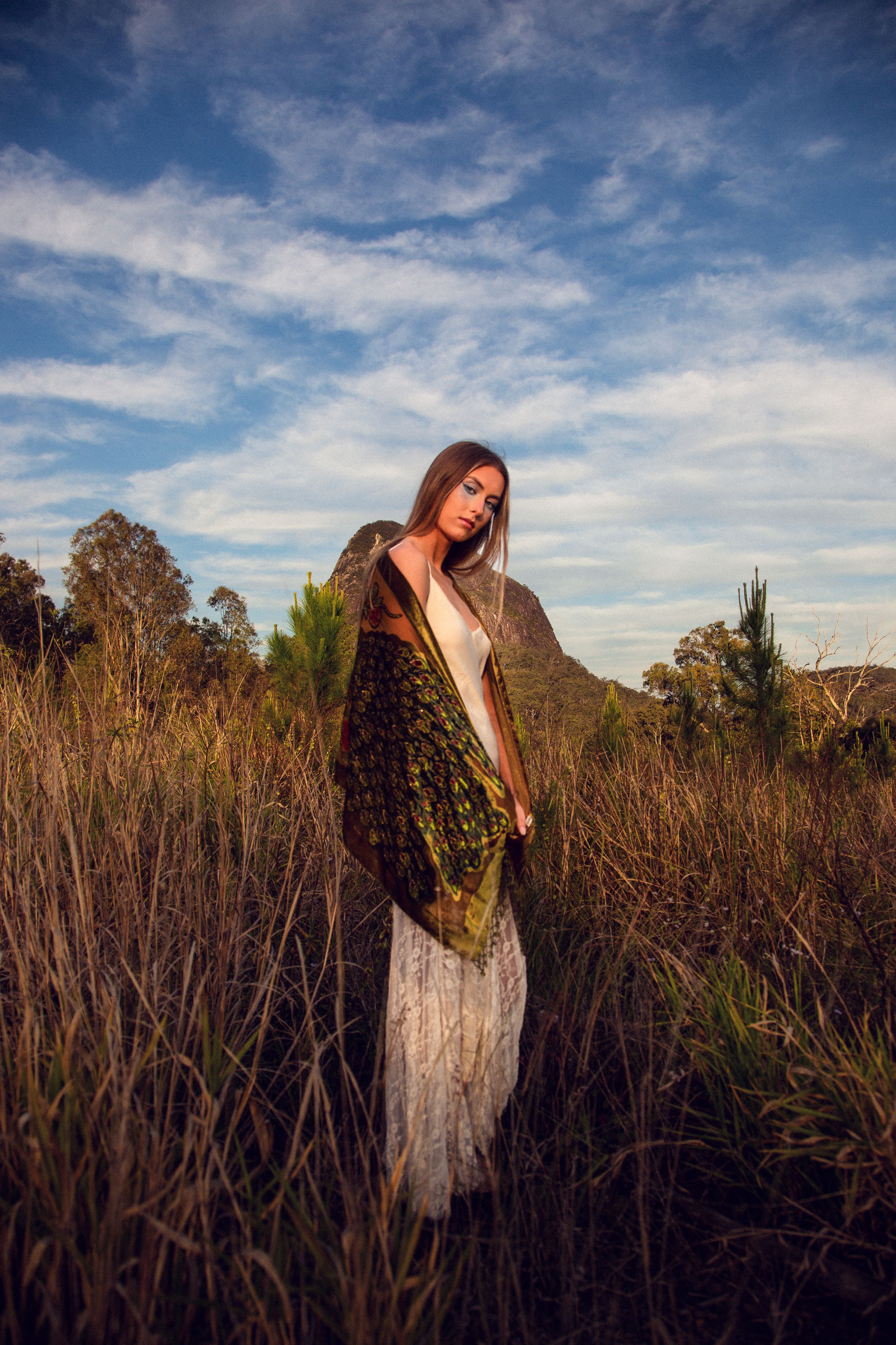 A figure stands in a field of tall grass at sunset wearing a flowing white dress and patterned shawl against a dramatic sky.