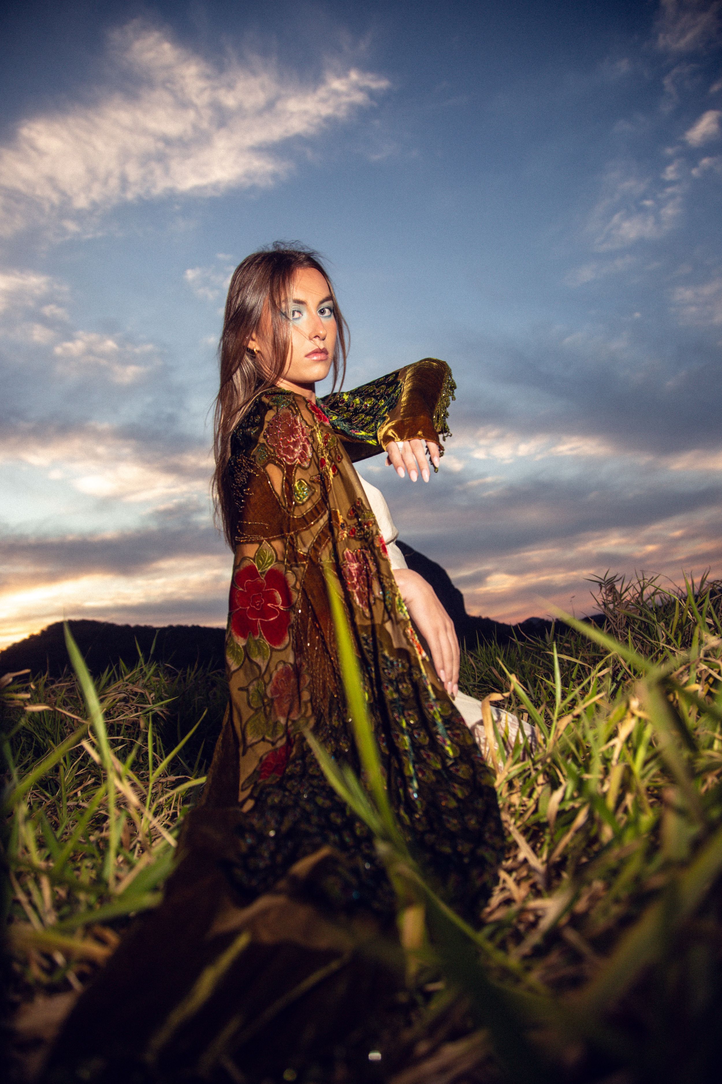 A woman in a vintage dress poses dramatically in a grassy field at sunset with dramatic cloudy sky.