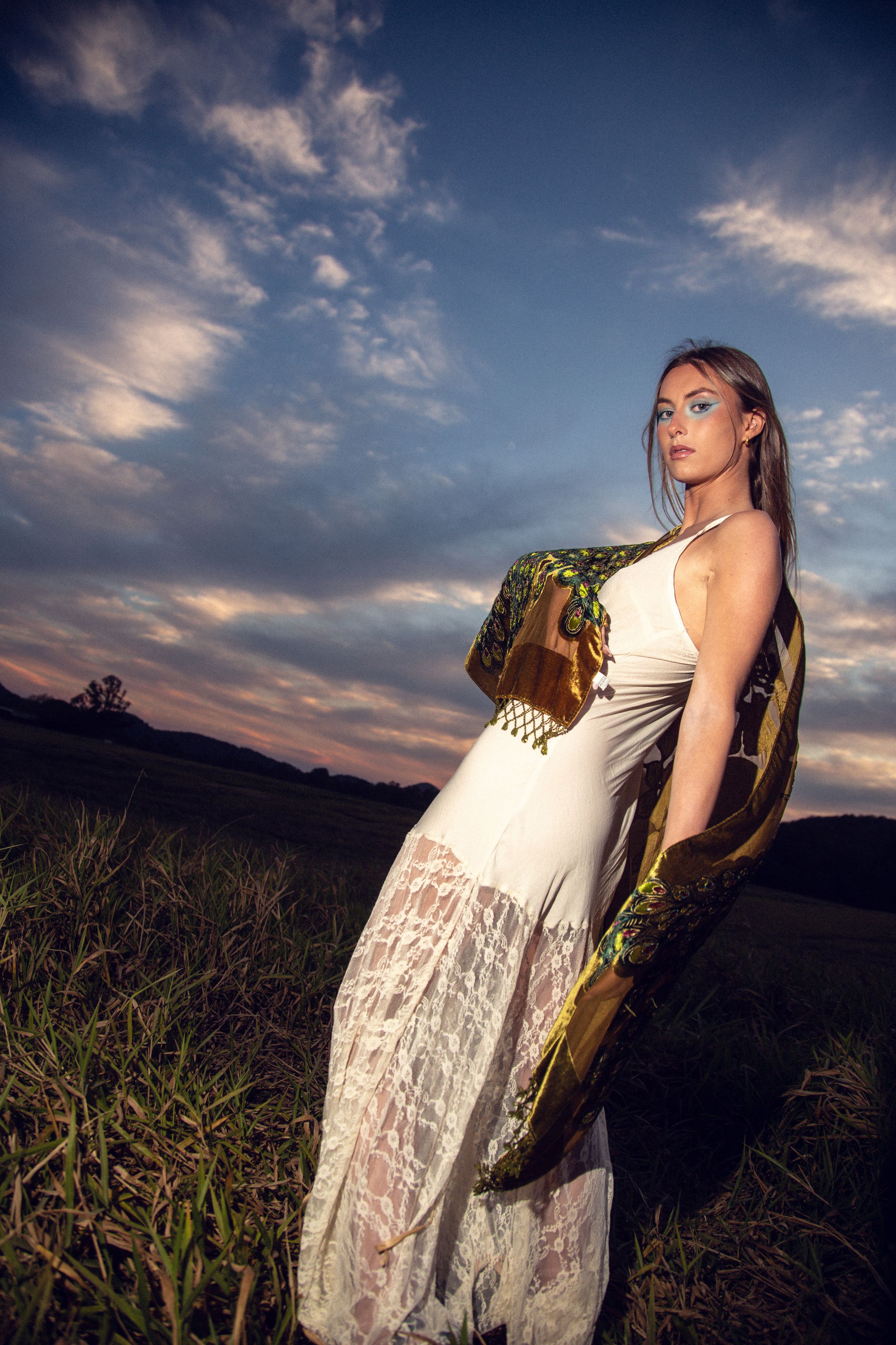 A figure in a flowing white lace dress poses dramatically against a sunset sky with dramatic clouds.