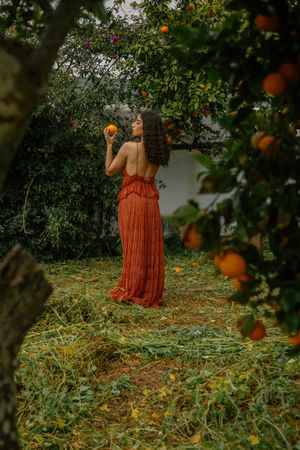 A figure in a flowing orange dress stands among citrus trees in a lush garden setting.