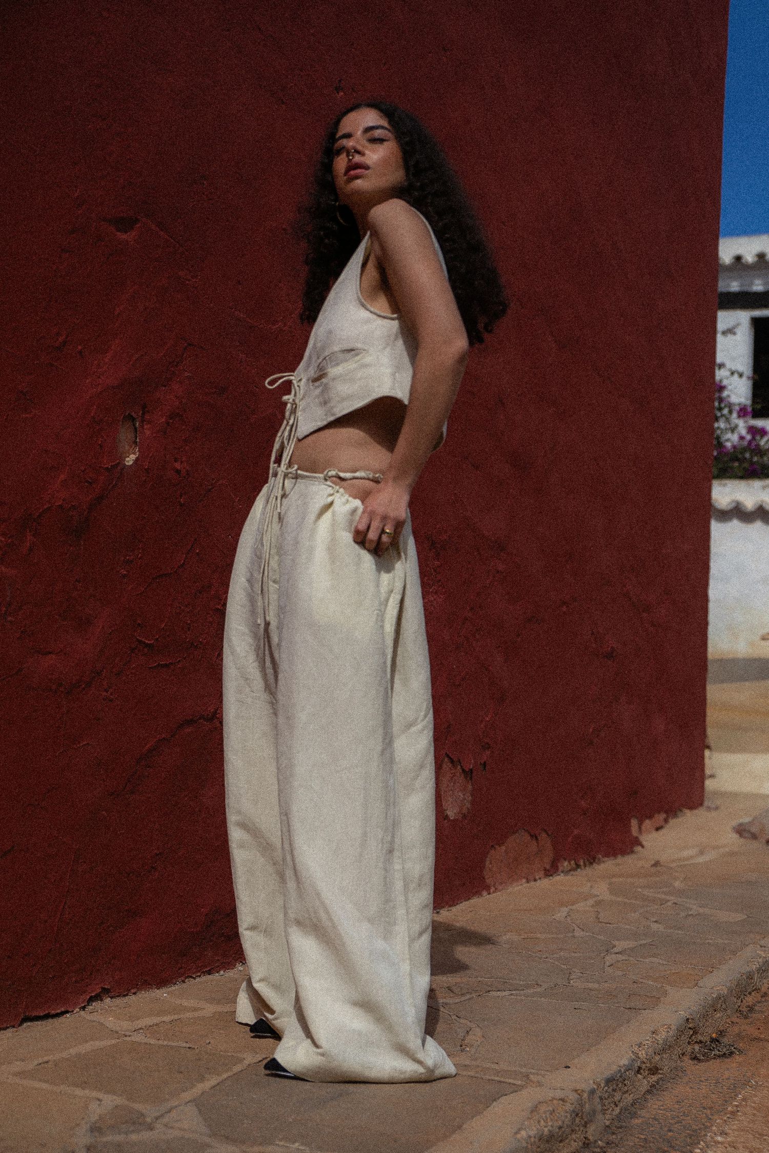 Woman in cream-colored linen crop top and wide-leg pants poses against a deep red wall in natural lighting.