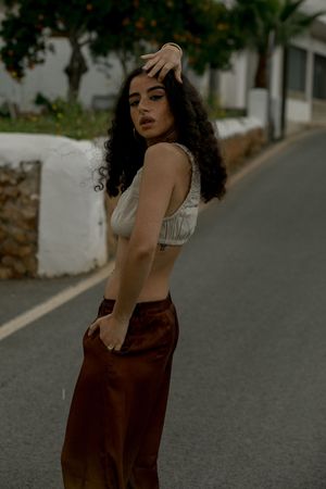 A person in a white crop top and burgundy skirt poses on a street with curly hair and dramatic makeup.