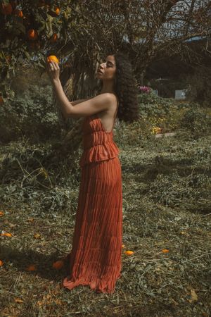 A moody fashion portrait in a garden features a rust-colored pleated maxi dress and reaching for citrus fruit.