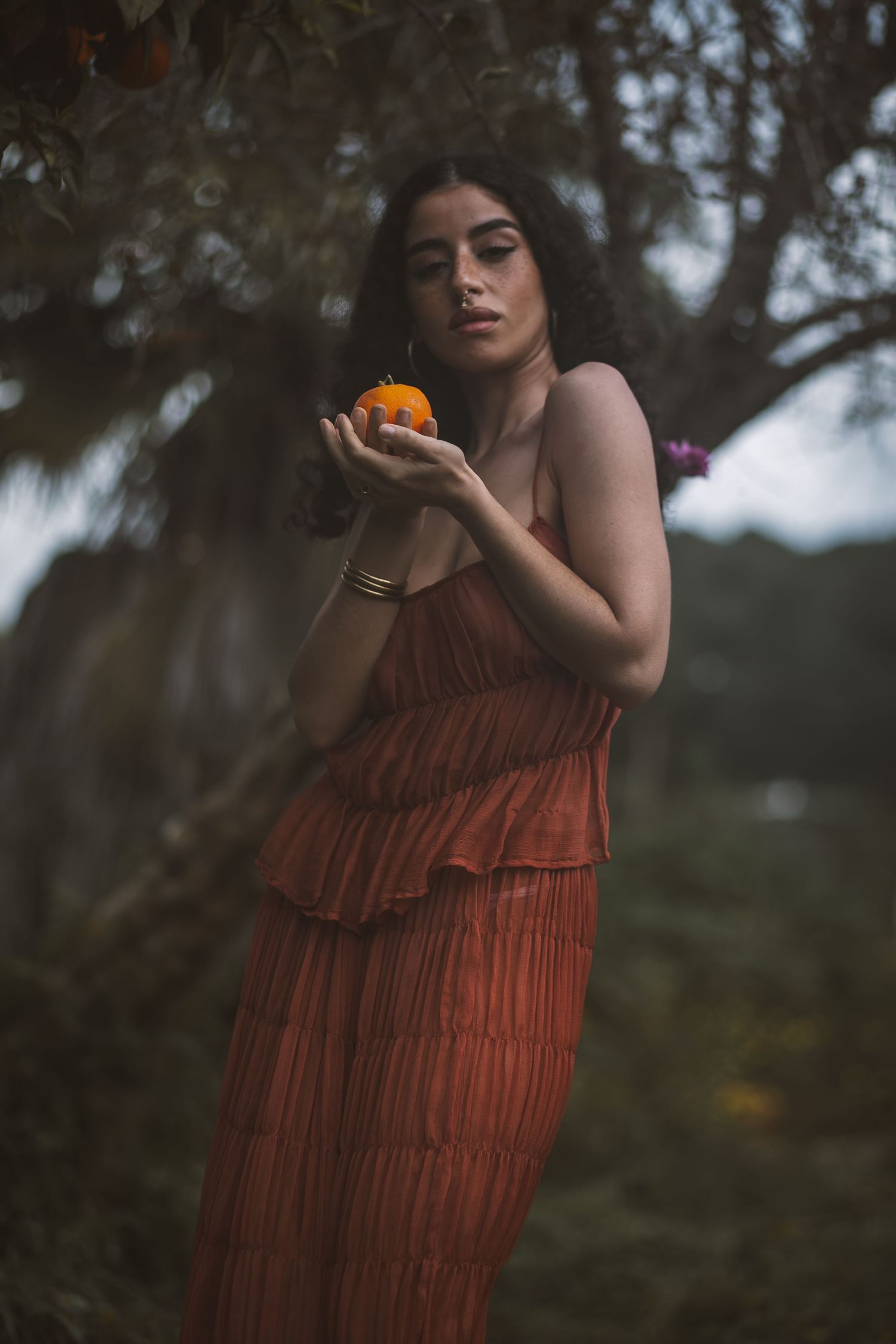 Artistic portrait in a garden shows a tiered rust-colored dress and marigold flower against dark foliage.