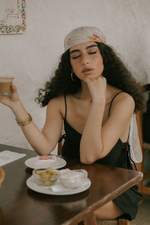 Artistic cafe scene with head scarf and black top, sitting at table with snacks and drinks.