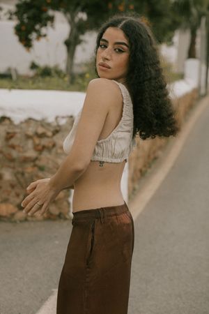 Fashion portrait with natural curls wearing a cropped white top and brown pants against a stone wall.