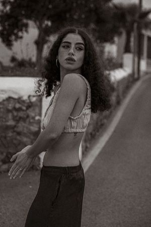Black and white portrait of someone with curly hair wearing a crop top and dark pants on a street.