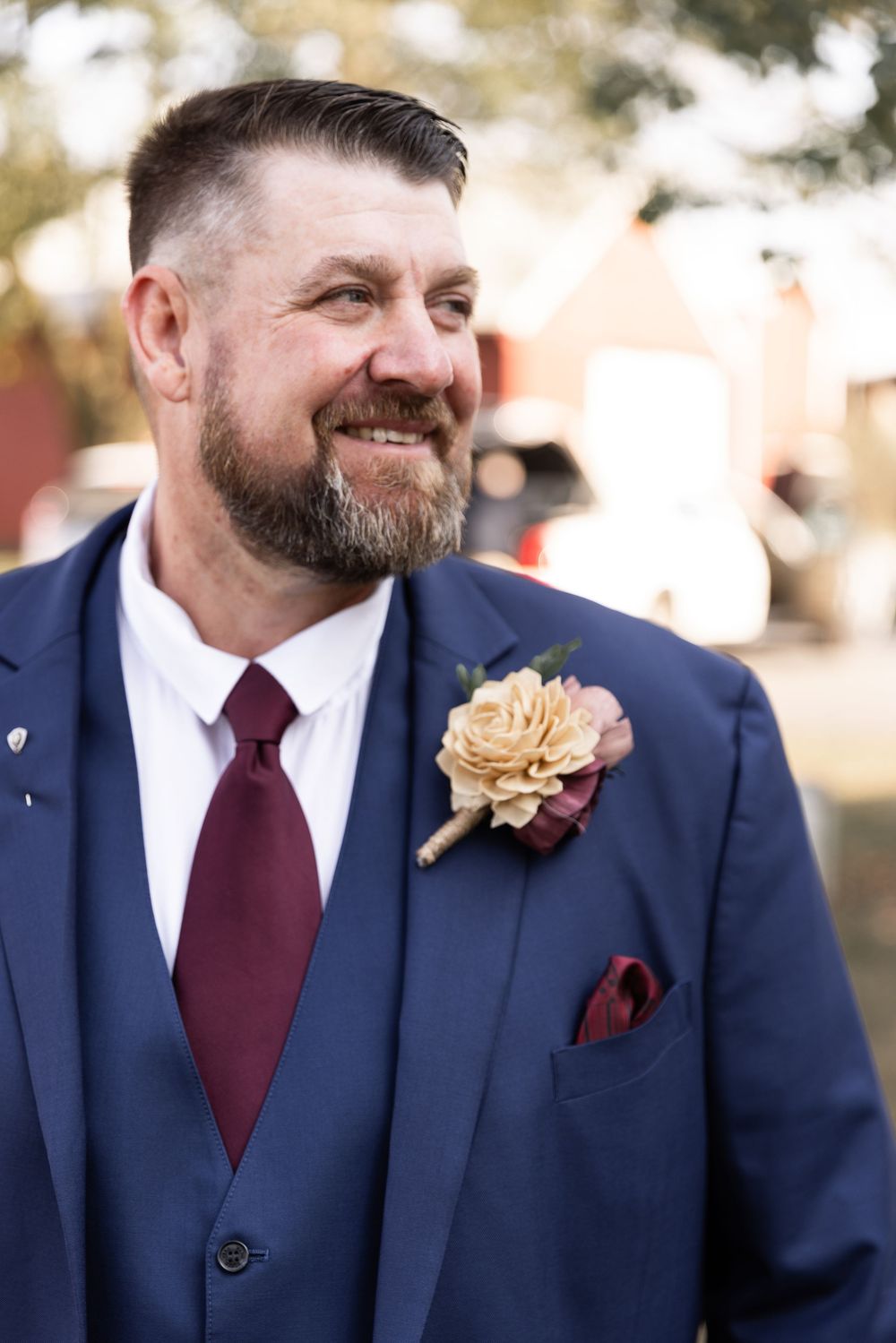 A groom wearing a navy suit and burgundy tie smiles while wearing a boutonniere at an outdoor wedding ceremony.