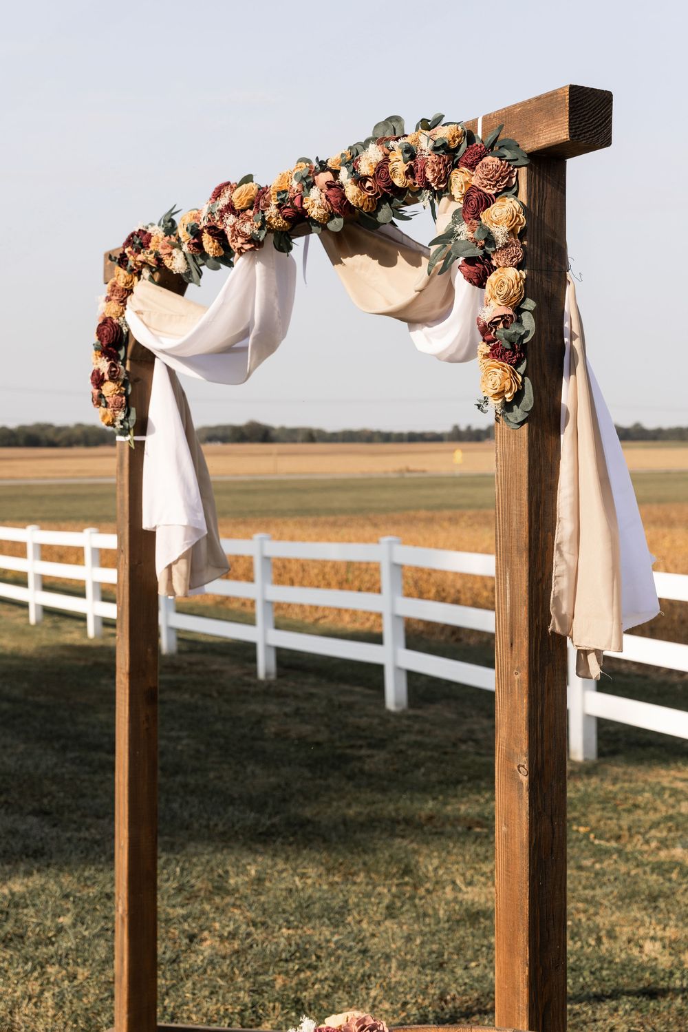 Rustic wooden wedding arch decorated with dried flowers and white fabric draping overlooking a white fence and field.