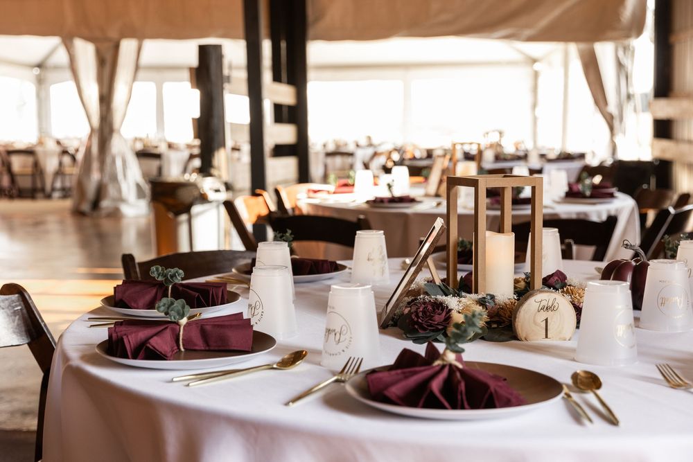 Elegant wedding reception table settings with burgundy napkins and silver place settings in a bright banquet hall.
