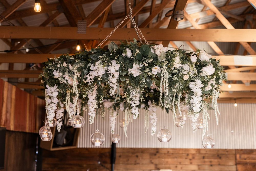 A rustic floral chandelier with cascading white flowers and greenery hangs from wooden barn rafters.