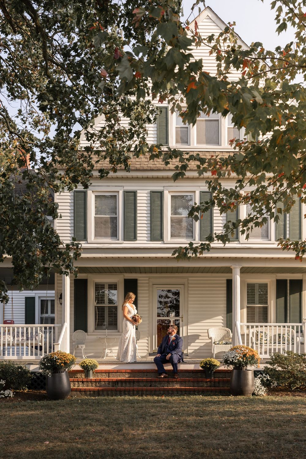 Classic white farmhouse with green shutters and wraparound porch surrounded by autumn trees.