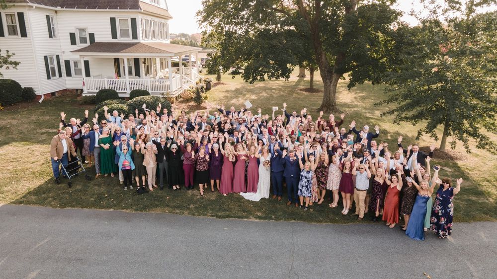 Large group of wedding guests pose together for panoramic photo in front of white colonial style house.